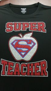 A black shirt with an embroidered apple with a Superman logo inside and the text “Super Teacher”